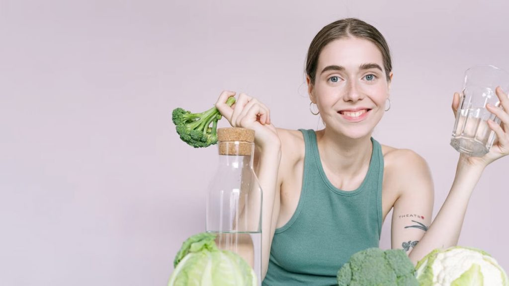 Woman in tank top smiling with broccoli, cabbage, and glass of water on table.