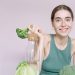 Woman in tank top smiling with broccoli, cabbage, and glass of water on table.