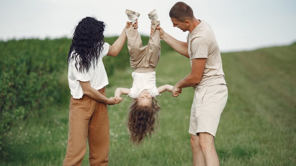 A joyful family enjoys outdoor play, lifting their child upside down in a green field.