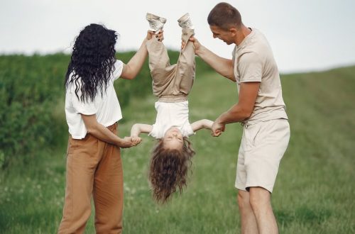 A joyful family enjoys outdoor play, lifting their child upside down in a green field.